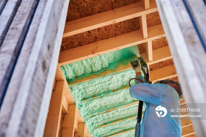 Male builder insulating wooden frame house. Man worker spraying polyurethane foam inside of future cottage, using plural component gun. Construction and insulation concept.