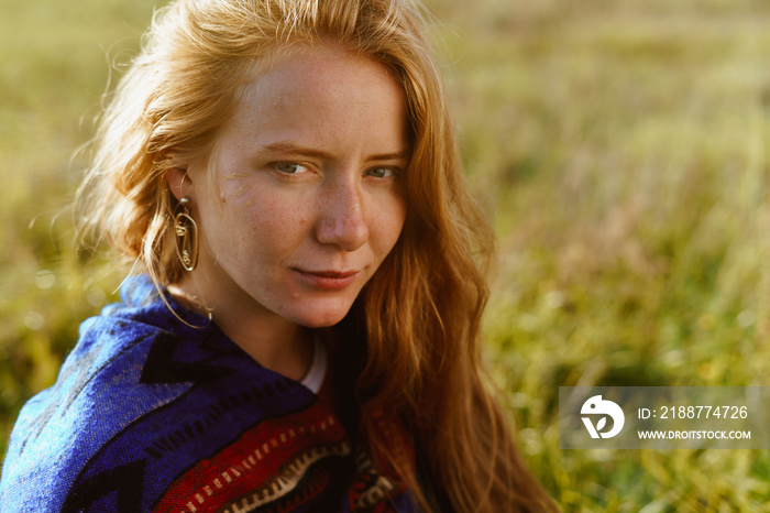 Meadow grass behind the back of a beautiful girl with long red hair and freckles on the face affectionate look of beautiful eyes without makeup and ethno cape on the shoulders. High quality photo