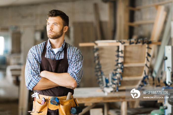 Waist-up portrait of confident bearded carpenter with arms crossed standing in spacious workshop and looking away