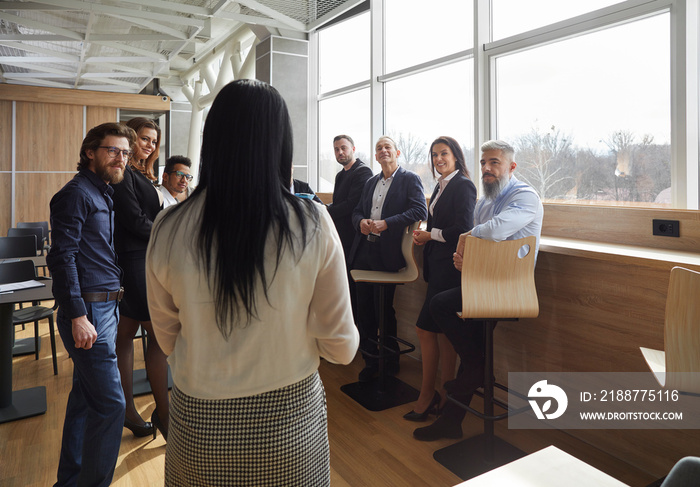 Team of diverse people having educational workshop or work meeting in office. Group of young and senior workers listening to woman who is a business coach or a corporate manager. Back view from behind