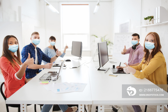 health, safety and pandemic concept - creative team wearing protective medical masks for protection from virus disease with computers showing thumbs up at office