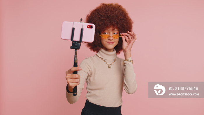 Young teenager child girl kid blogger taking selfie on smartphone selfie stick, communicating video call talking online with subscribers. Little funny children isolated on studio pink background