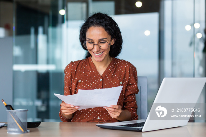 Young happy and successful businesswoman in glasses working with documents inside office, Hispanic woman with laptop looking at bills and contracts, financier with curly hair using laptop.