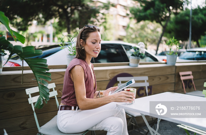 Smiling beautiful female influencer dressed in trendy wear checking mail and messages on digital tablet drinking cocktail on terrace,woman using touchpad connected to 4G for reading news online