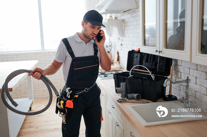 Busy handyman talk on phone in kitchen. He hold hose. Toolbox on desk. Guy stand at sink.