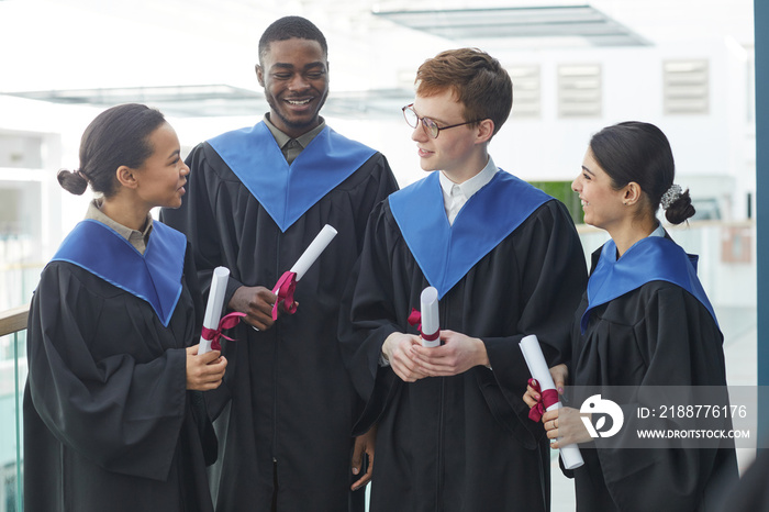 Waist up view at diverse group of young people wearing graduation gowns indoors in modern university interior and chatting