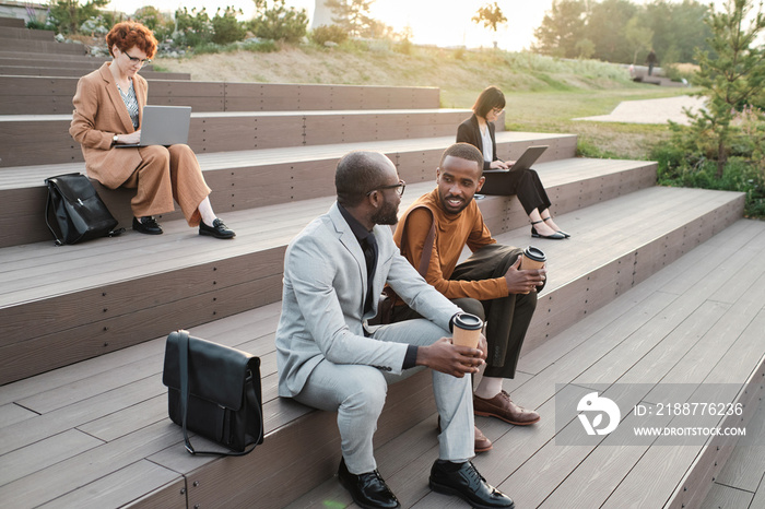 Horizontal high angle view shot of modern ethnically diverse men and women spending time outdoors in urban park working and having coffee break