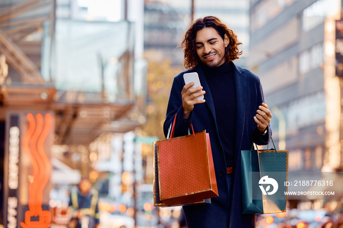 Happy Latin American man with shopping bags using cell phone on street.