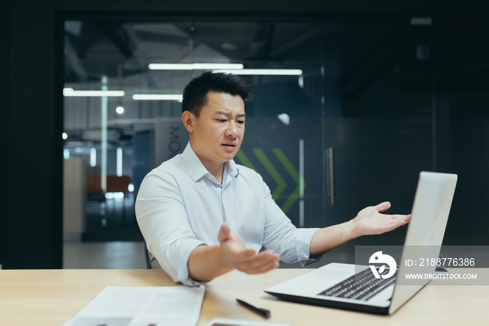 A young handsome man, a confused Asian businessman, sits at a laptop in the office, talks on a video call, spreads his hands, shows that he does not understand. In a modern office.