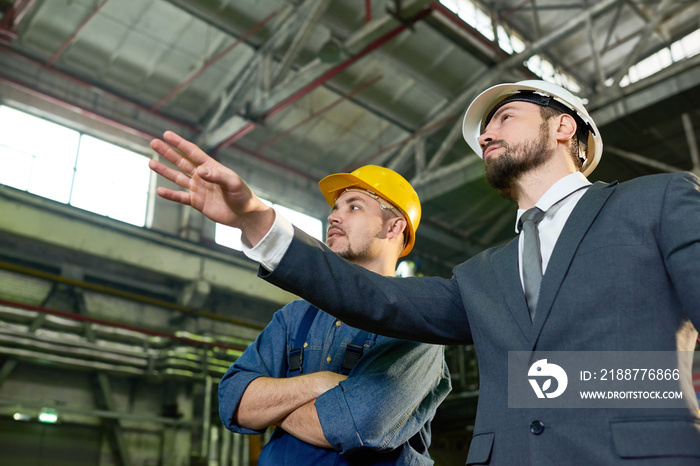 Low angle portrait of handsome businessman wearing hardhat pointing to something while discussing production with worker at modern plant, copy space