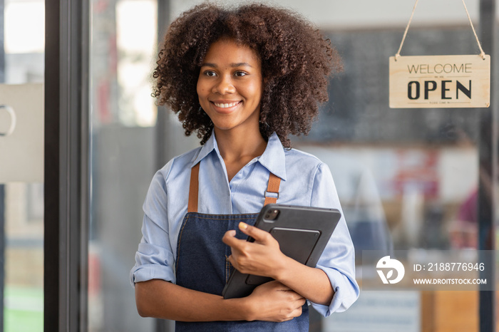 Successful african woman in apron standing coffee shop door. Happy small business owner holding tablet and working. Smiling portrait of SME entrepreneur seller business standing with copy space.