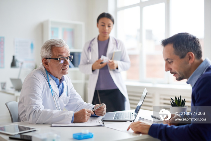 Senior male doctor in white coat talking to the patient at the table they discussing method of treatment with nurse in the background