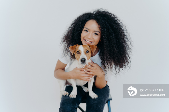 Smiling African American woman poses with domestic animal, looks gladfully at camera, cuddles dog, sits on chair, isolated over white background. Two family members. Homely relax. Forever together