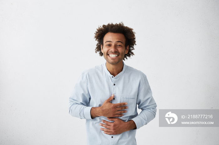 Positive attitude and genuine human emotions. Waist up portrait of cheerful joyful young African man holding hands on stomach while laughing out loud at something funny, not suppressing his feelings