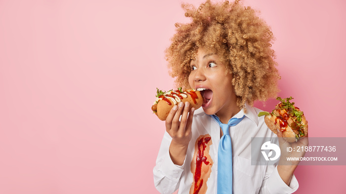 Photo of hungry curly woman eats hot dog and burger keeps mouth widely opened prefers fast food wears white formal shirt and tie isolated over pink background blank space for your advertisement