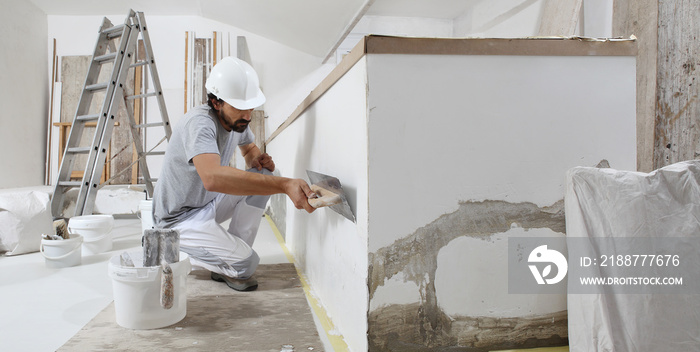 man plasterer construction worker at work, takes plaster from bucket and puts it on trowel to plastering the wall, wears helmet inside the building site of a house