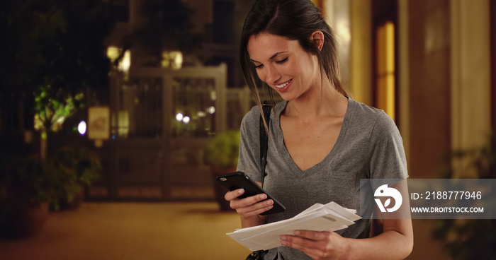 Woman with mail checking smartphone and walking offscreen in outdoor courtyard