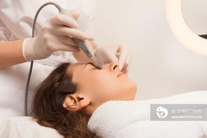 Cosmetologist is cleaning a womans face with the ultrasound spatula. White theme.