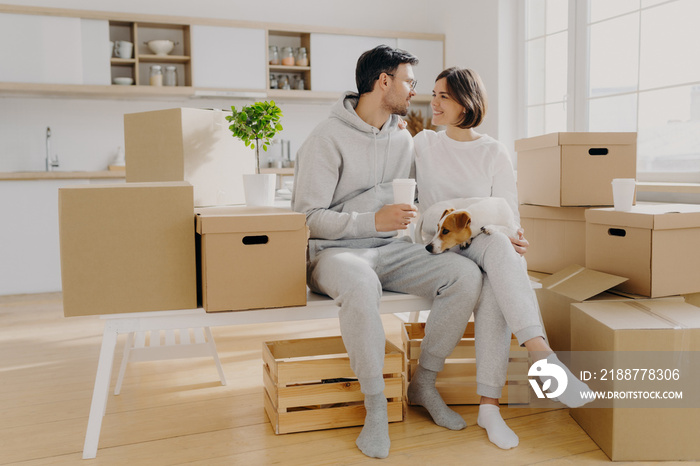 Lovely spouses sit near carton big boxes, drink coffee and talk about future plans, relocate in new apartment, pose with pedigree dog, poses in spacious kitchen, think about design or interior