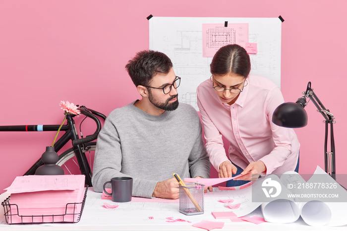 Two female male coworkers pose at office desk discuss ideas for future project study paper documents and stickers. Woman assistant help man to prepare construction sketches. Collaboration concept