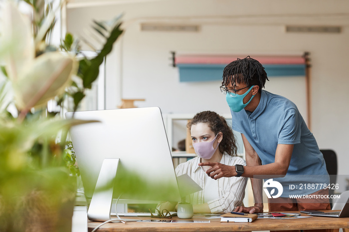 Portrait of two young photographers wearing masks while pointing at computer screen and reviewing pictures in office studio, copy space