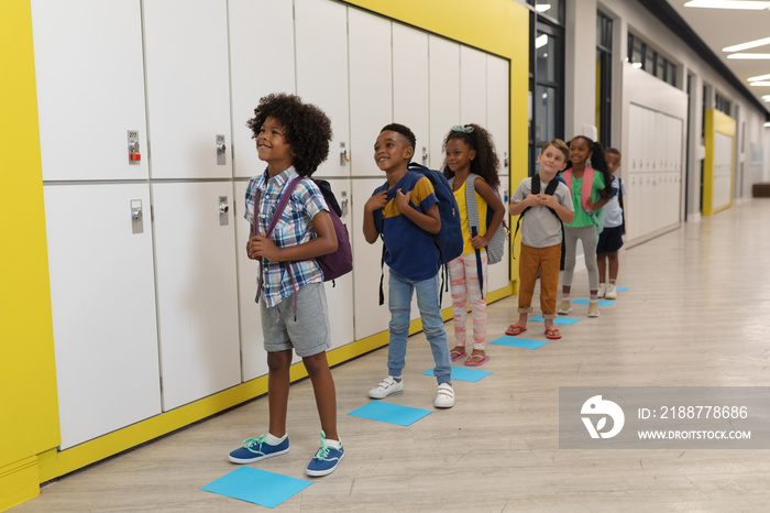 Full length of multiracial elementary school children with backpack standing by lockers in corridor