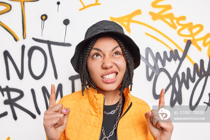 Beautiful ethnic hipster girl with cheeky expression clenches teeth points above demonstrates something against graffiti wall dressed in fashionable outfit. Modern gen z teen has cool outlook