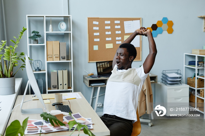 Portrait of exhausted young man stretching and yawning at office workplace