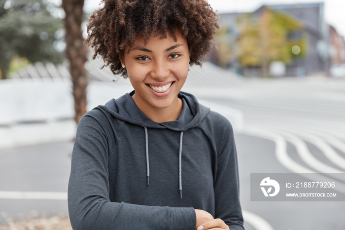 Outdoor lifestyle concept. Photo of pleasant looking happy cool teenager with Afro haircut, dressed in black oversized hoodie, spends free time on sportsground, enjoys youth, dreams about something