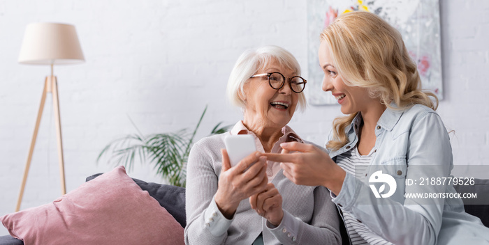 Cheerful woman pointing at smartphone in hands of senior mother, banner