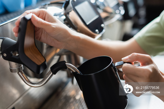 Cropped view of barista holding milk jug near coffee machine with steam wand in cafeteria.