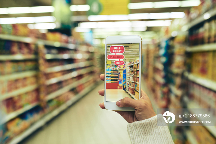 Close-up shot of young woman doing shopping at hypermarket with help of augmented reality app, blurred background