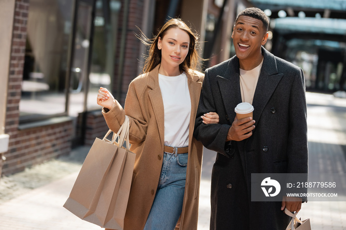 funny and cheerful african american man looking at camera near young girlfriend with shopping bags.