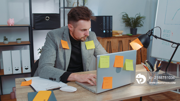 Tired exhausted businessman in suit working on laptop at office with many sticker task inscriptions. Freelancer man busy occupation feels sad boredom overworked multitasking. Panic attack, deadline