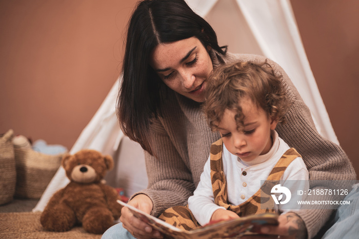 a mother reading a book to her little son in the bedroom before going to sleep. caucasian mom telling stories sitting on the floor.