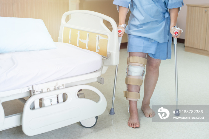 Asian woman patient with knee brace and walking stick in hospital ward after ligament surgery.