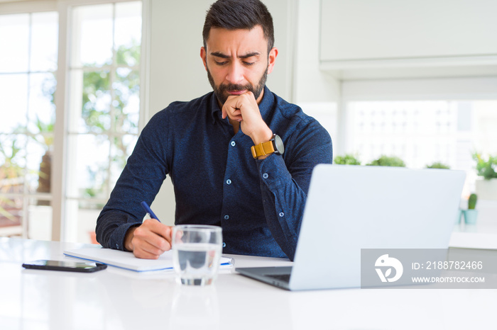 Handsome hispanic man working using computer and writing on a paper serious face thinking about question, very confused idea