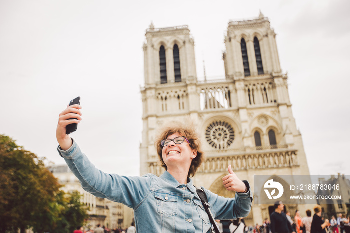 Tourist in Paris making funny selfie near Notre Dame Cathedral. Beautiful young Caucasian tourist woman with backpack in Paris making funny selfie hand holding phone, photo near Notre Dame Cathedral