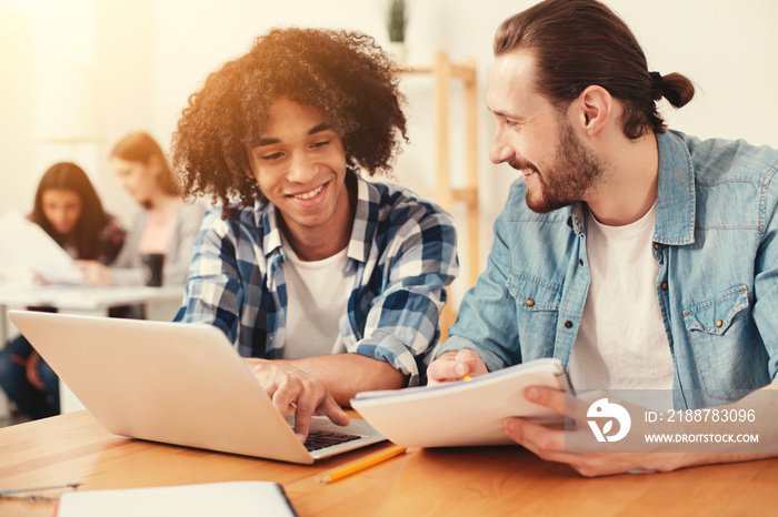 Understanding. Cheerful friendly men smiling while looking at the screen of a laptop and reading notes in a notebook