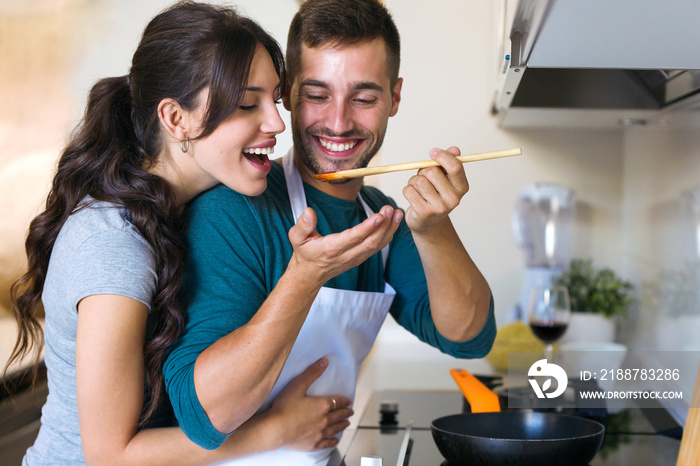 Handsome young man giving his wife to try the food he is preparing in the kitchen at home.