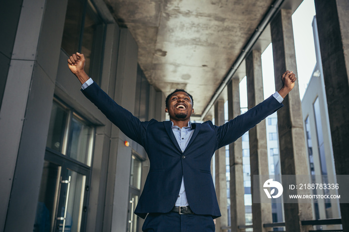 excited african american businessman rejoices in victory looking at camera while standing outside office building. Happy male freelancer in formal suit, shouts success, winner, win or good profit