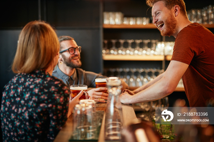 Cheerful friends leaning on bar counter, drinking beer and chatting with bartender. Night out.