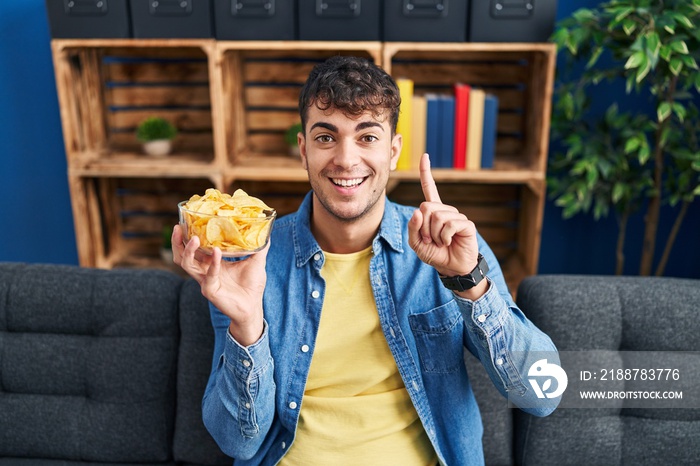 Young hispanic man holding potato chips surprised with an idea or question pointing finger with happy face, number one