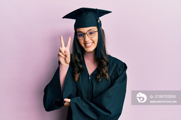 Young hispanic woman wearing graduation cap and ceremony robe smiling with happy face winking at the camera doing victory sign. number two.