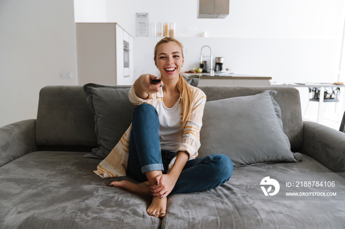 Happy blonde woman watching tv with remote control while sitting on couch