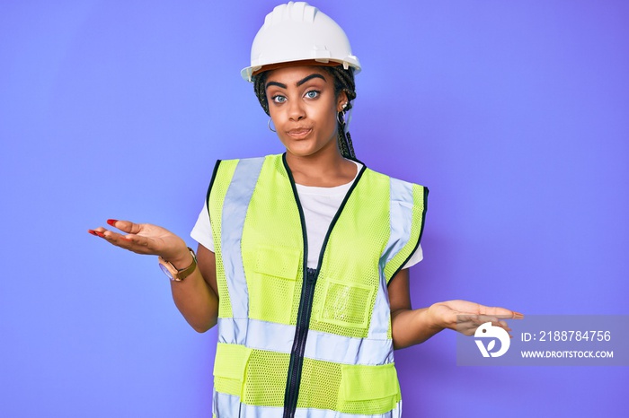 Young african american woman with braids wearing safety helmet and reflective jacket clueless and confused expression with arms and hands raised. doubt concept.