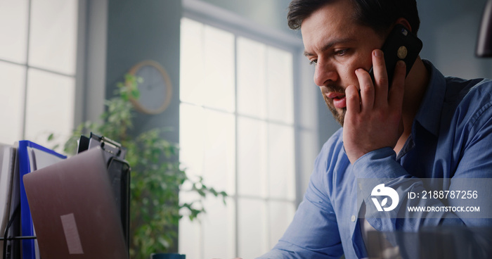 Nervous frustrated man sitting at desk talking on the phone, looking upset with conversation. Young businessman working from home office, chatting on mobile, having trouble.