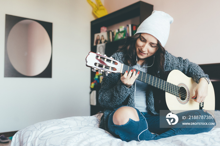 Smiling young woman playing guitar while sitting on bed at home