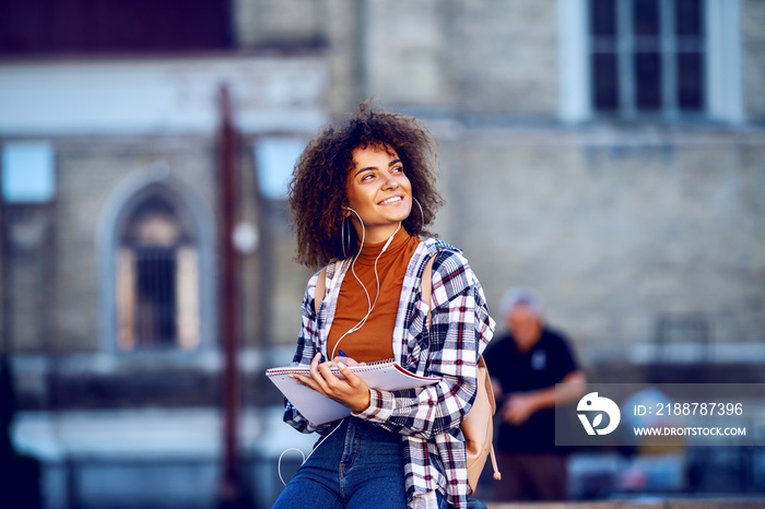 Smiling cute mixed race student with curly hair standing outdoors with earphones in ears, writing in notebook and looking away.