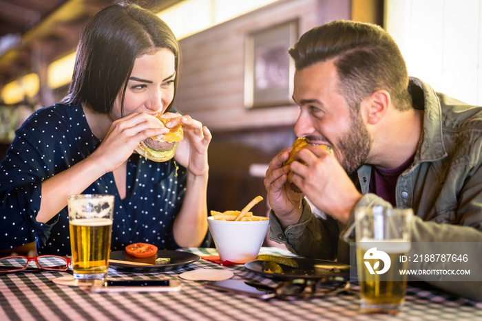 Happy couple having fun eating burger at restaurant pub fast food - Young people enjoying moment at indoor diner location - Relationship concept with focus on girl face with warm vivid filter